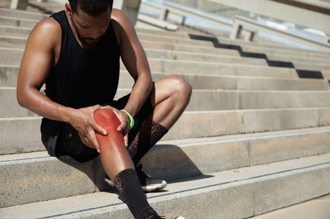 Cropped portrait of dark-skinned jogger in training socks and running shoes having sprain, clutching his injured knee, massaging it with both hands after intense workout in urban surroundings